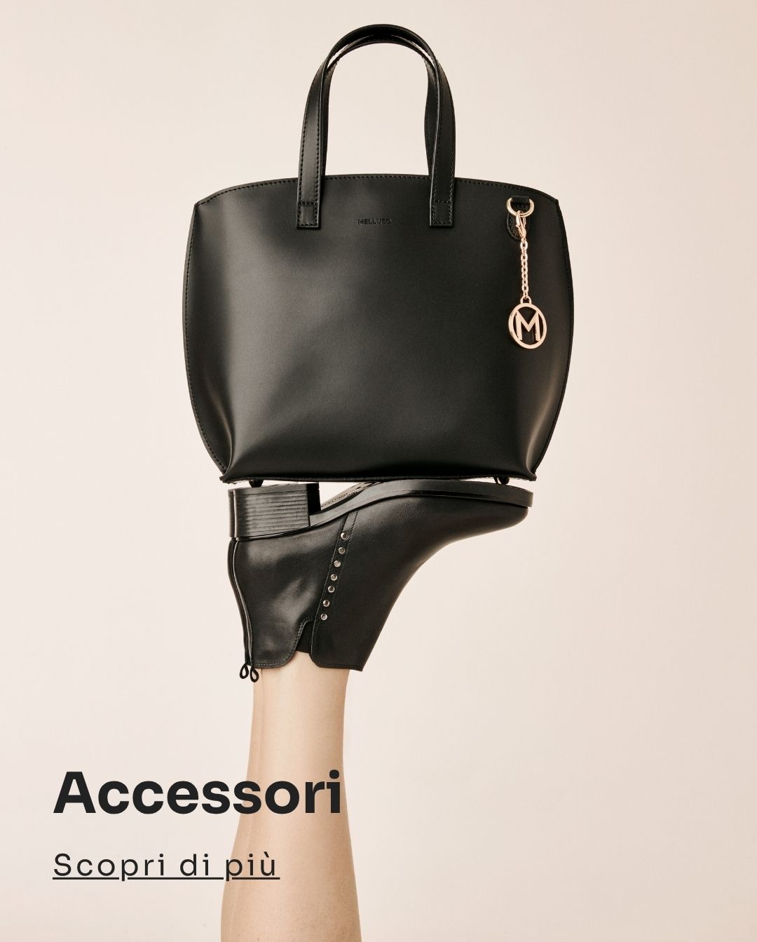 Black leather tote handbag with gold 'M' charm balanced on a black ankle boot worn on a raised leg against a minimalist beige background