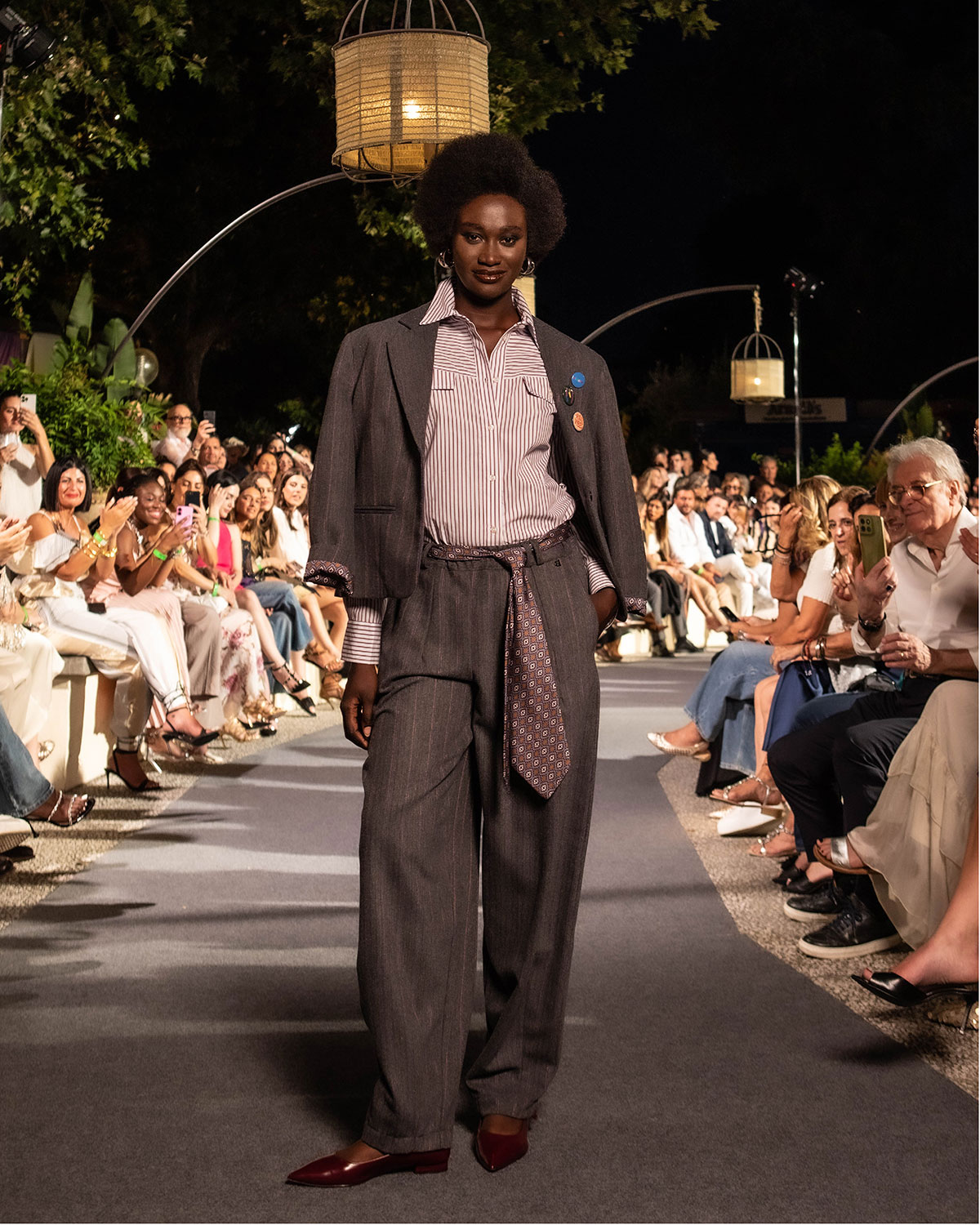 Model in oversized pinstripe suit, striped shirt and patterned tie walks outdoor evening runway with seated audience