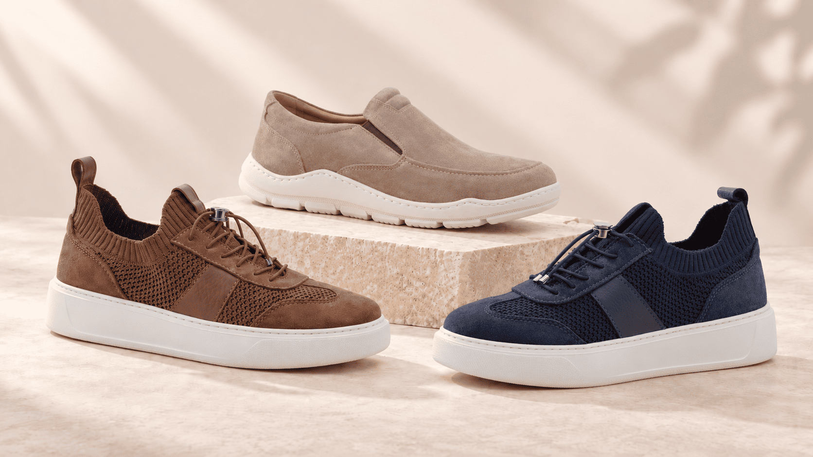 Three men's casual knit sneakers (navy, brown, beige) with white soles displayed on a stone pedestal against a neutral backdrop