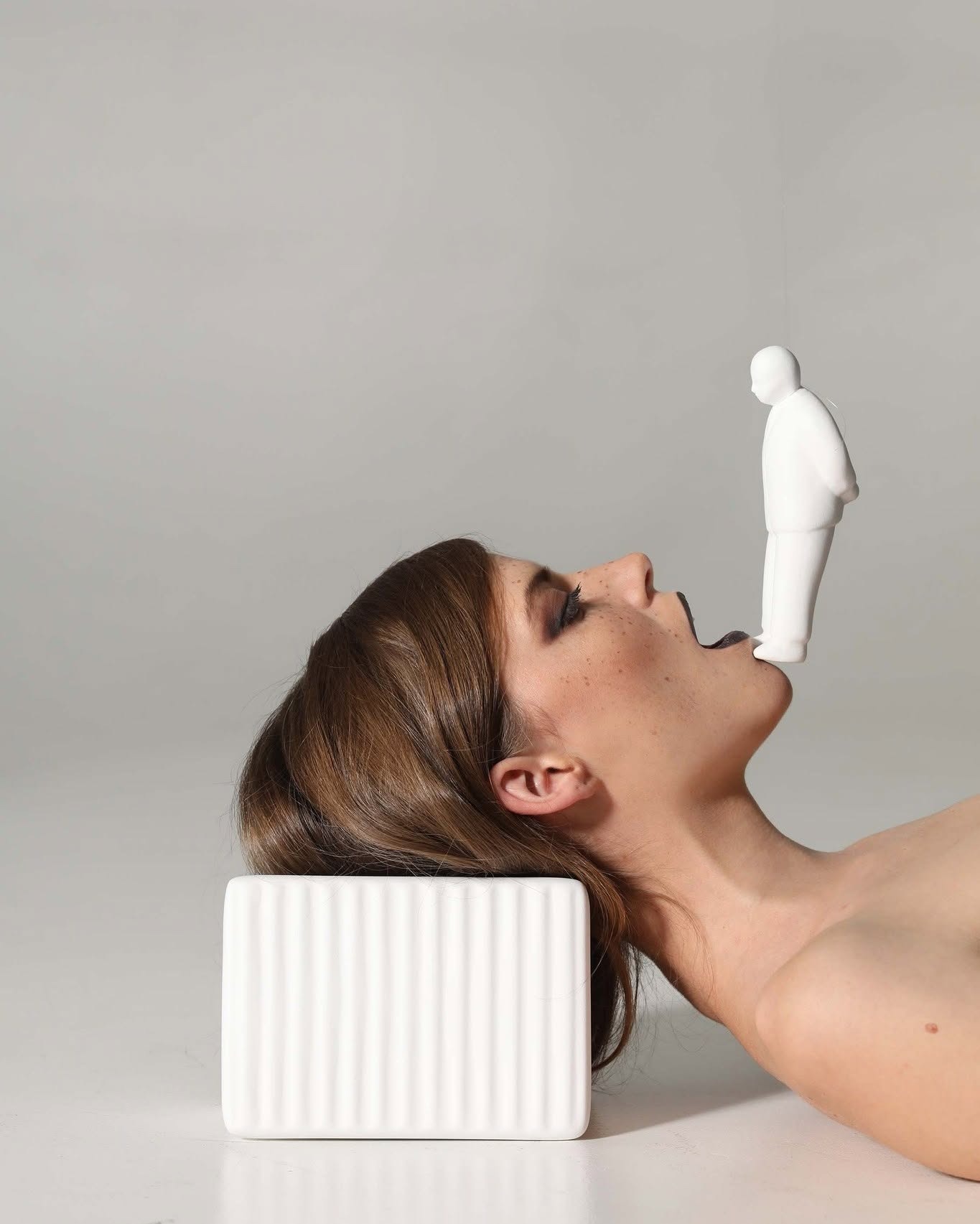 Surreal studio photo of woman lying with head on a white block, mouth open as a small white figurine balances above it