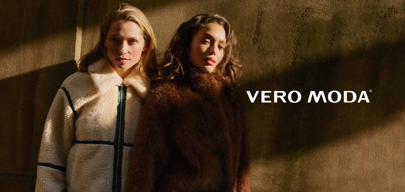 Due donne in piedi vicine, indossano giacche invernali testurizzate: una crema con dettagli neri, l'altra marrone scuro, davanti a un muro in ombra.