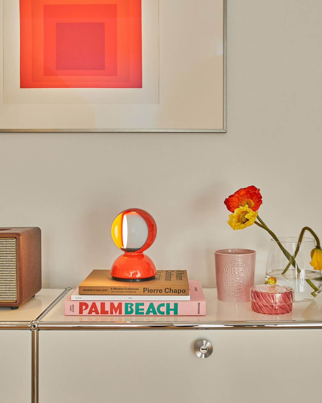 Minimalist sideboard with orange orb lamp on stacked 'Palm Beach' books, framed geometric wall art and vase of red-yellow poppies
