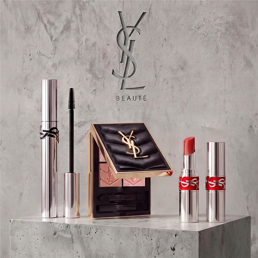 Un mascara argento YSL, una palette di ombretti nera trapuntata e un rossetto corallo esposti su un piedistallo di cemento.