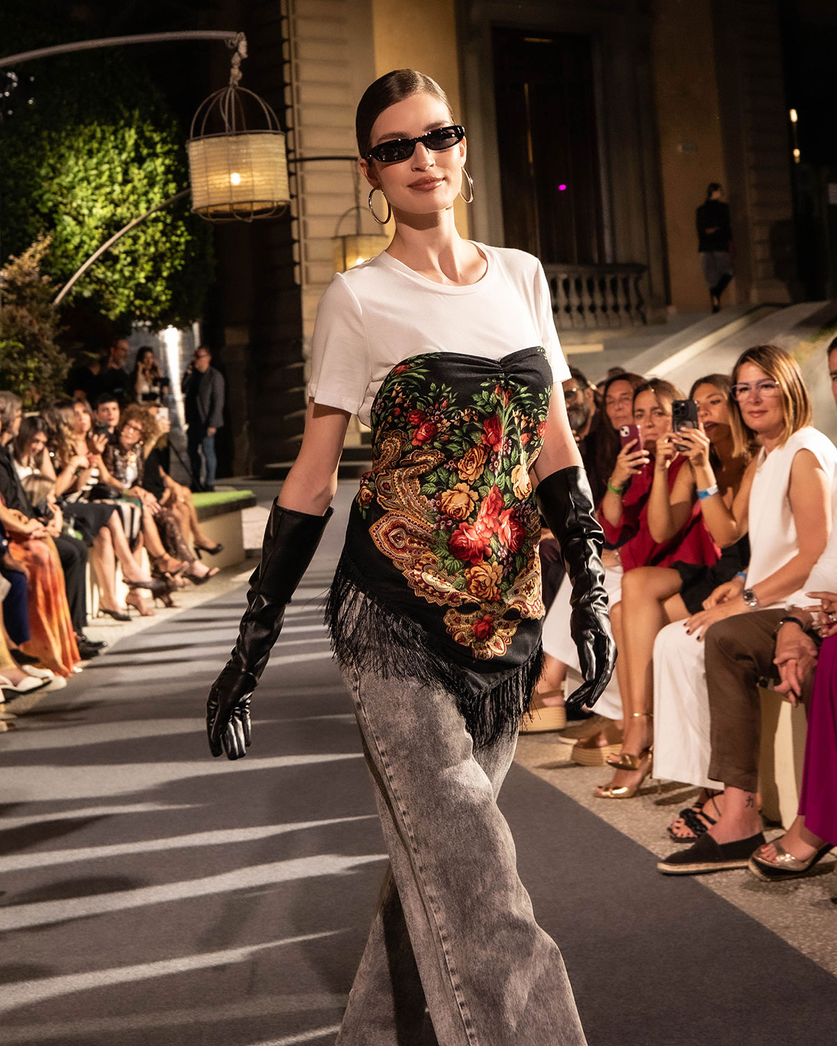 Model on fashion runway wearing tee with floral scarf-style top, long black leather gloves, sunglasses, audience watching.