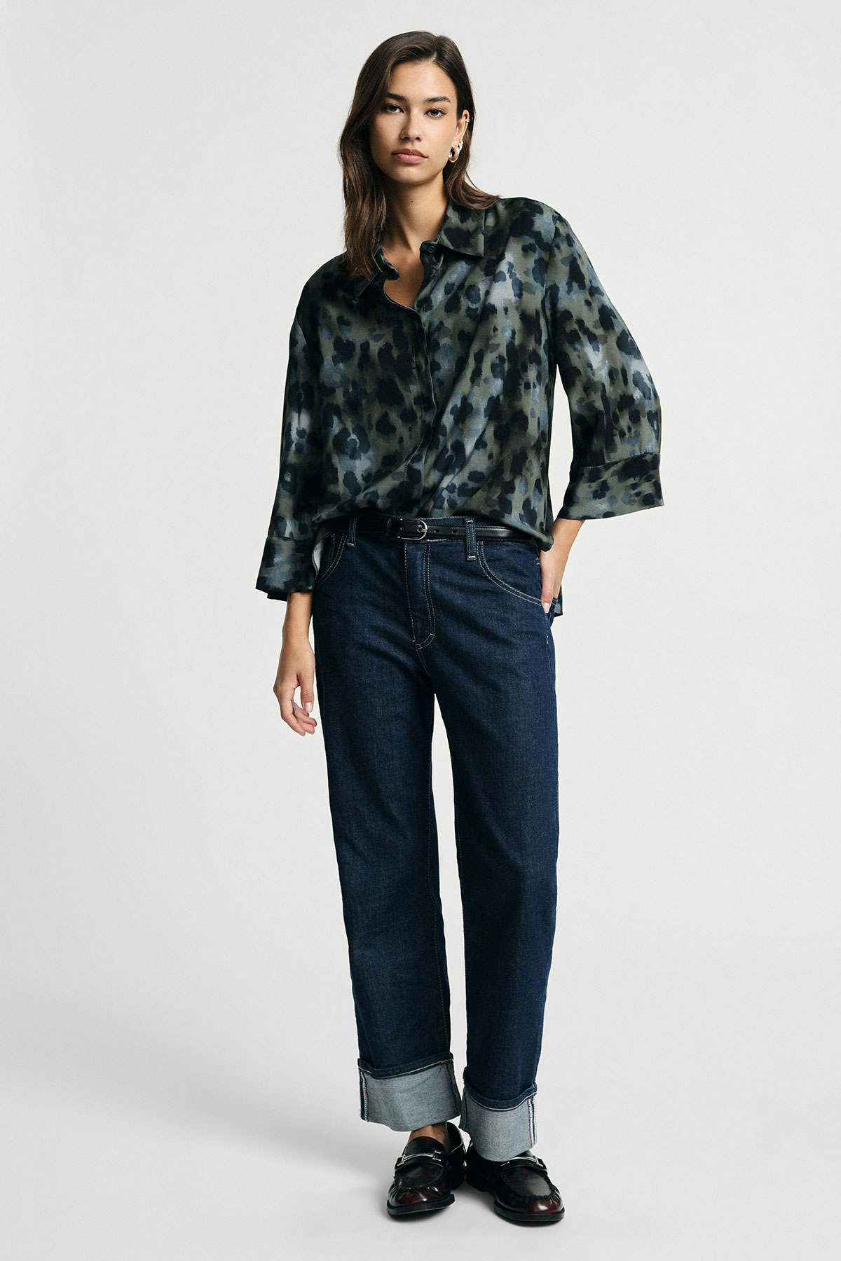 Full-length shot of a woman in a green patterned silk blouse, cuffed dark blue jeans and black loafers
