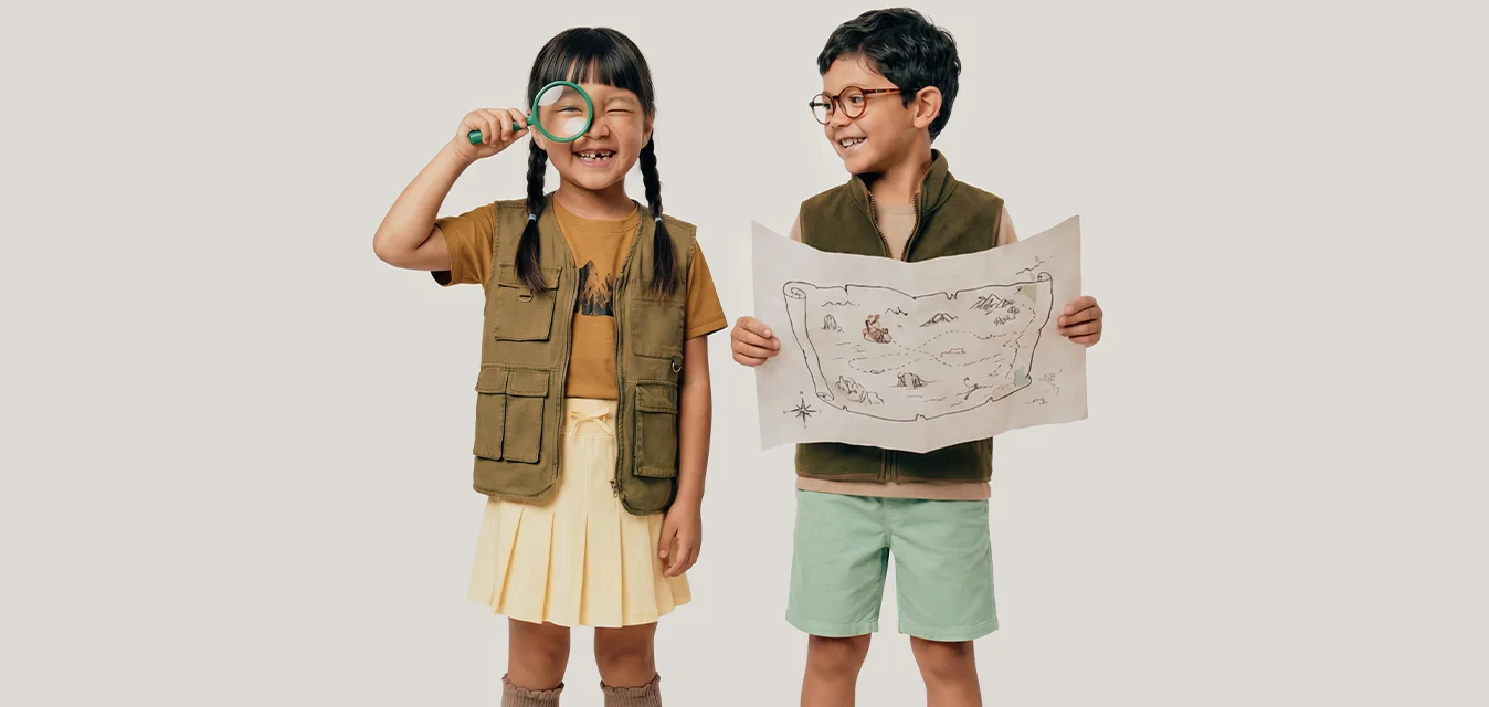 Due bambini vestiti da esploratori, uno tiene una lente d'ingrandimento e l'altro una mappa del tesoro, entrambi sorridenti.
