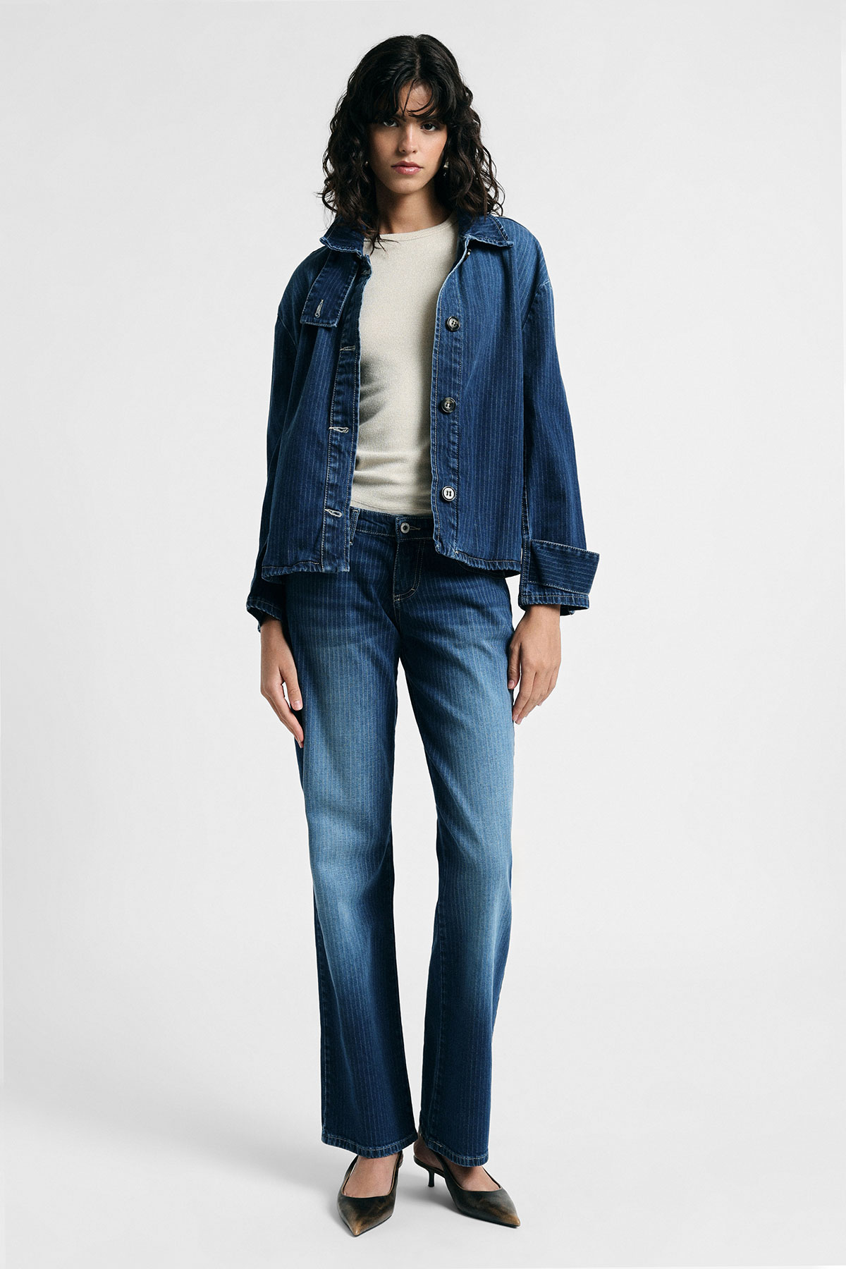 Woman modelling blue denim jacket and matching straight-leg jeans with cream top and black heels on plain white background