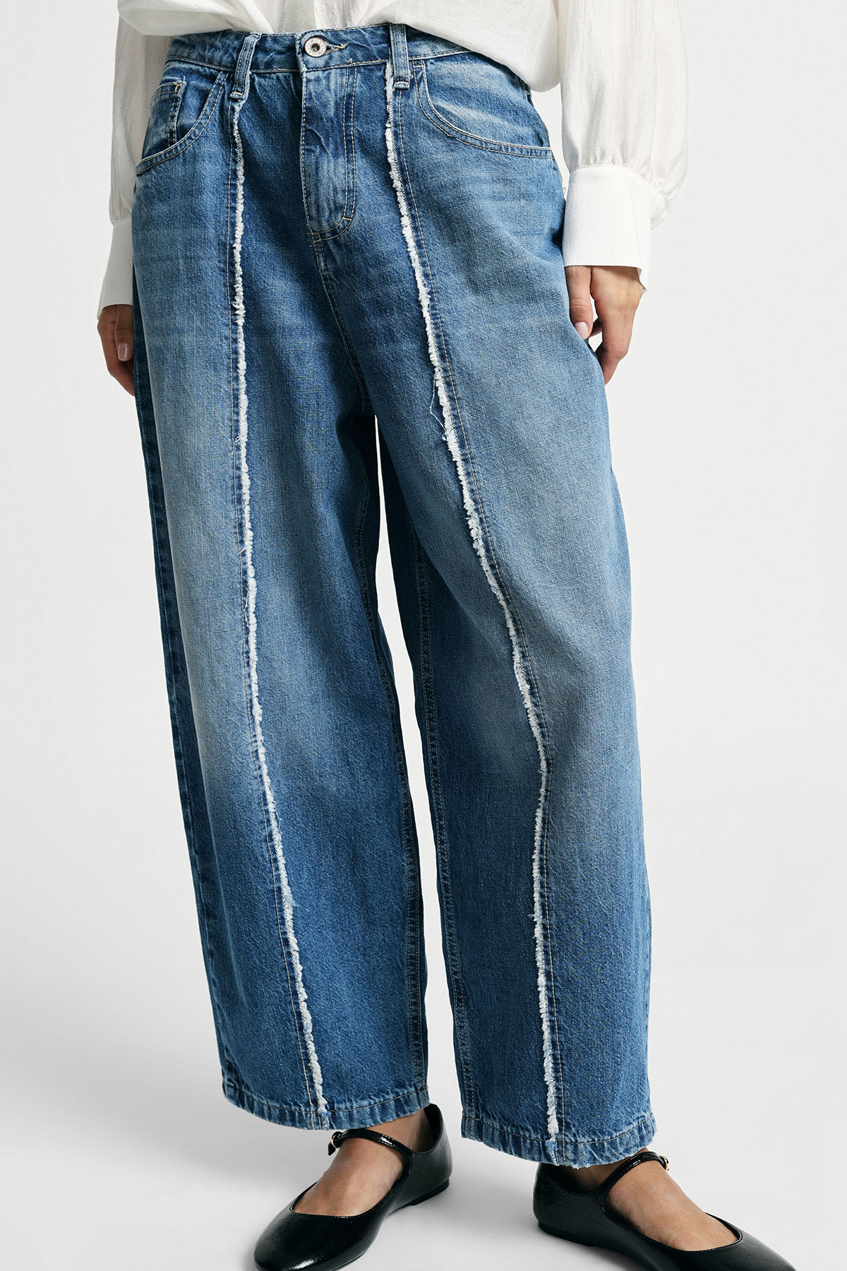 Wide-leg blue denim jeans with vertical raw exposed seams, worn with a white blouse and black Mary Jane flats