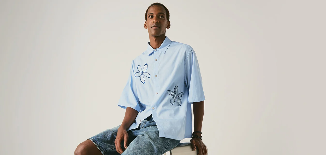Giovane uomo seduto su uno sgabello con una camicia azzurro chiaro con stampe floreali e pantaloncini di jeans su uno sfondo chiaro semplice.