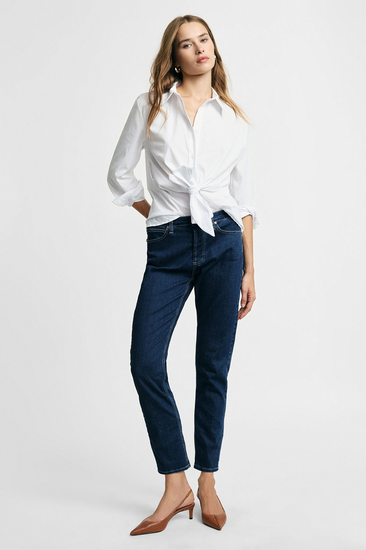 Model wearing white tie-front shirt, dark blue tapered jeans and tan kitten-heel shoes on plain background
