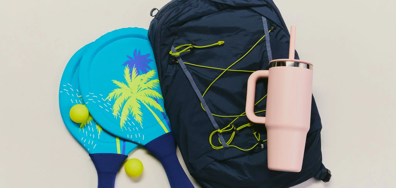 Due racchette da spiaggia blu con palline gialle, uno zaino scuro, una tazza da viaggio rosa e auricolari verdi disposti su una superficie chiara.
