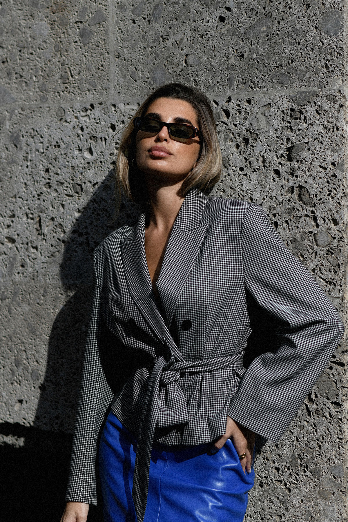 Woman in sunglasses and checked belted blazer with blue leather skirt leaning against textured concrete wall