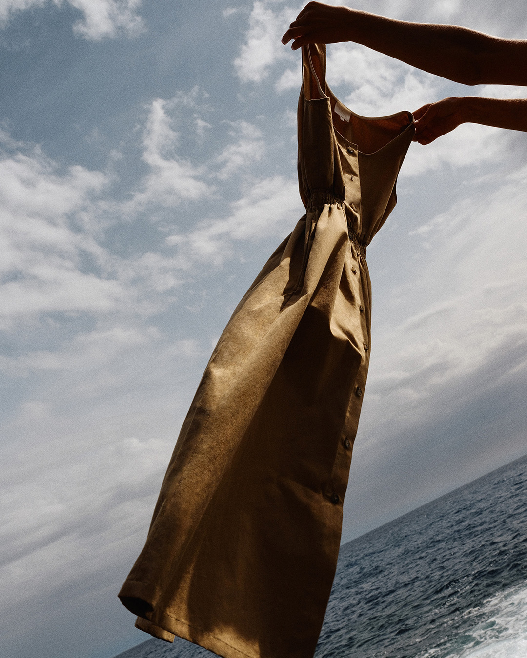 Sleeveless brown button-front midi dress held up by hands against a cloudy sky and sea backdrop