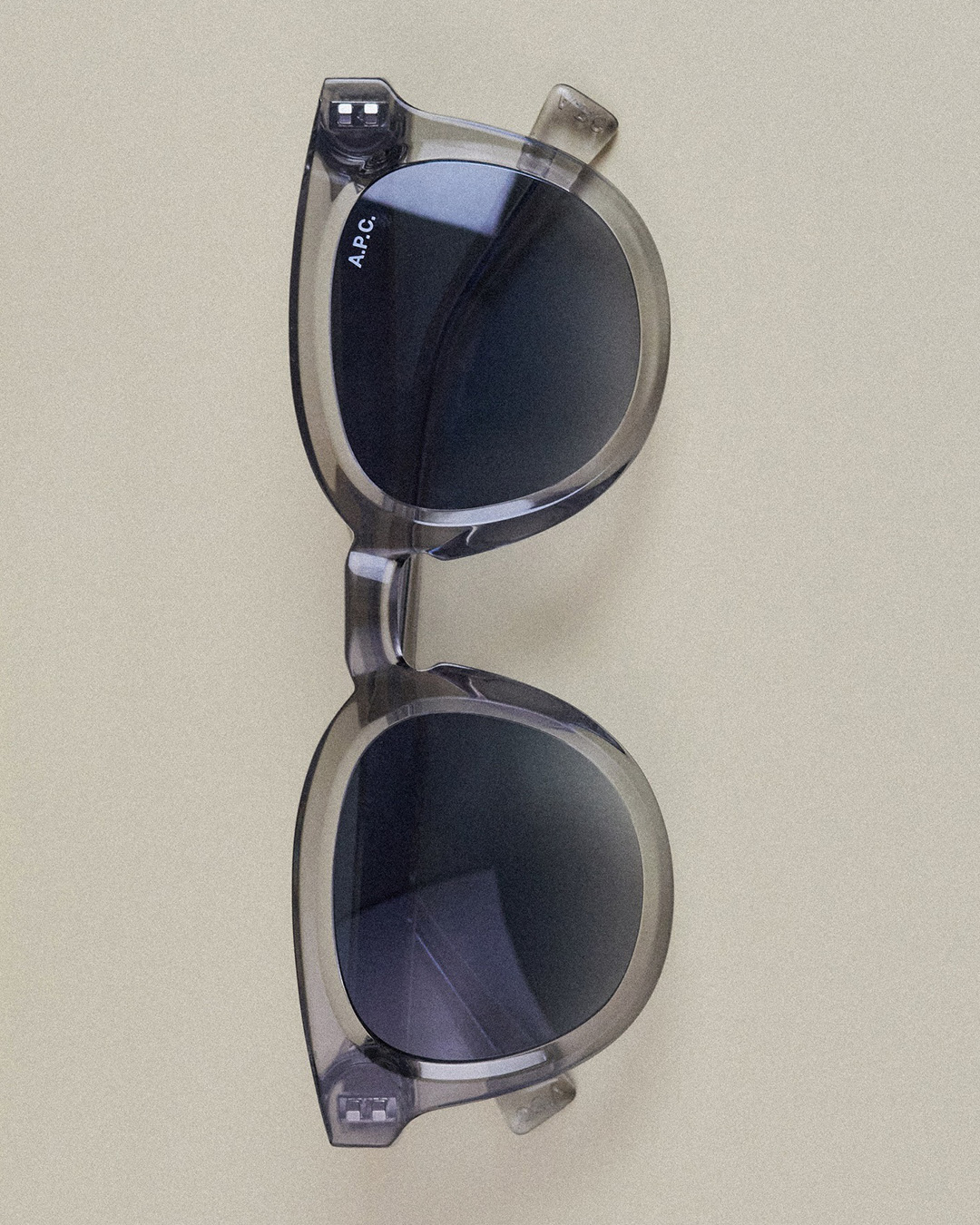Top-down view of translucent grey A.P.C. round sunglasses with dark lenses on a beige background
