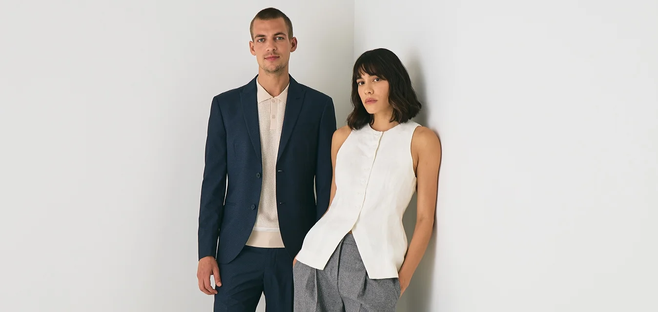 Uomo in abito blu navy e sneakers bianche, donna in top bianco senza maniche e pantaloni larghi grigi, in piedi in un angolo bianco minimal.