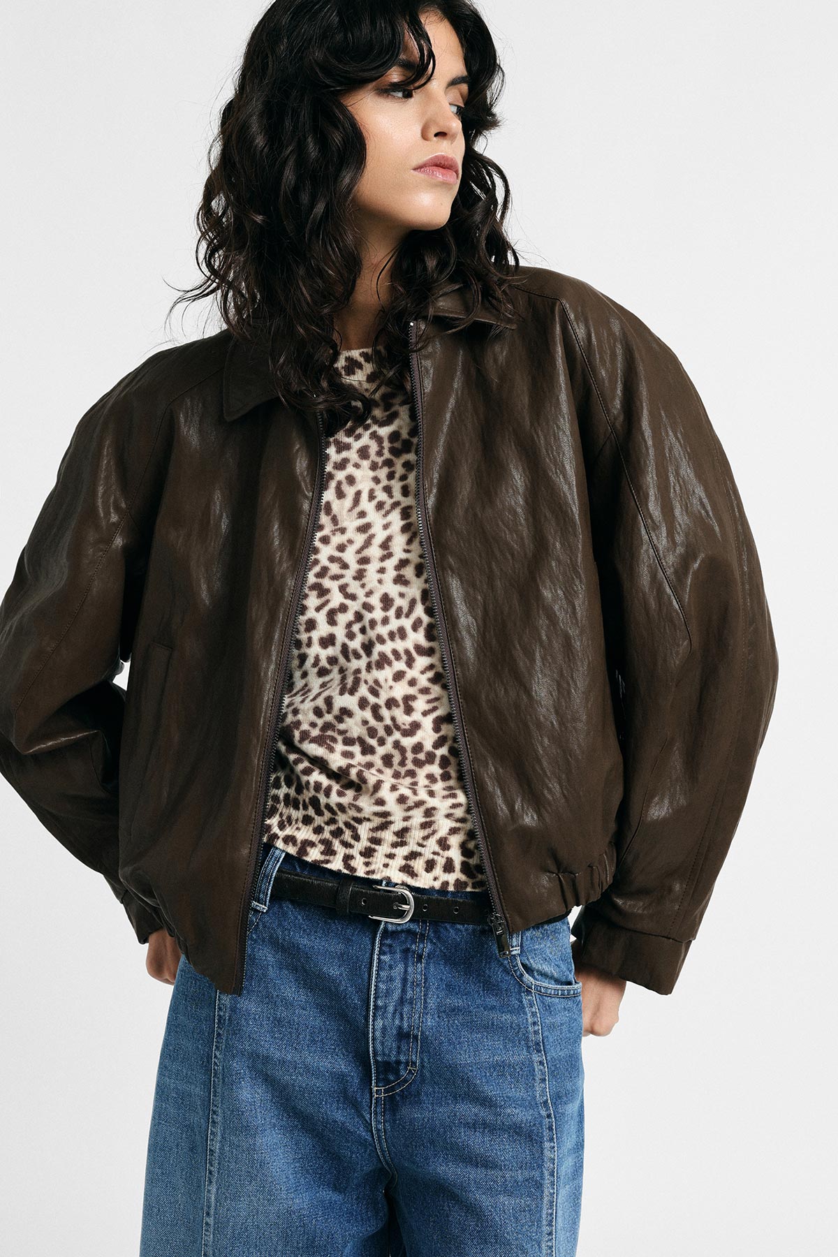 Young woman in brown leather bomber jacket, leopard-print top and high-waist blue jeans, hands in pockets, looking left