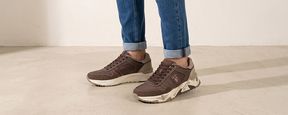 Brown leather-look men's trainers with chunky cream patterned soles, worn with cuffed blue jeans on a pale floor