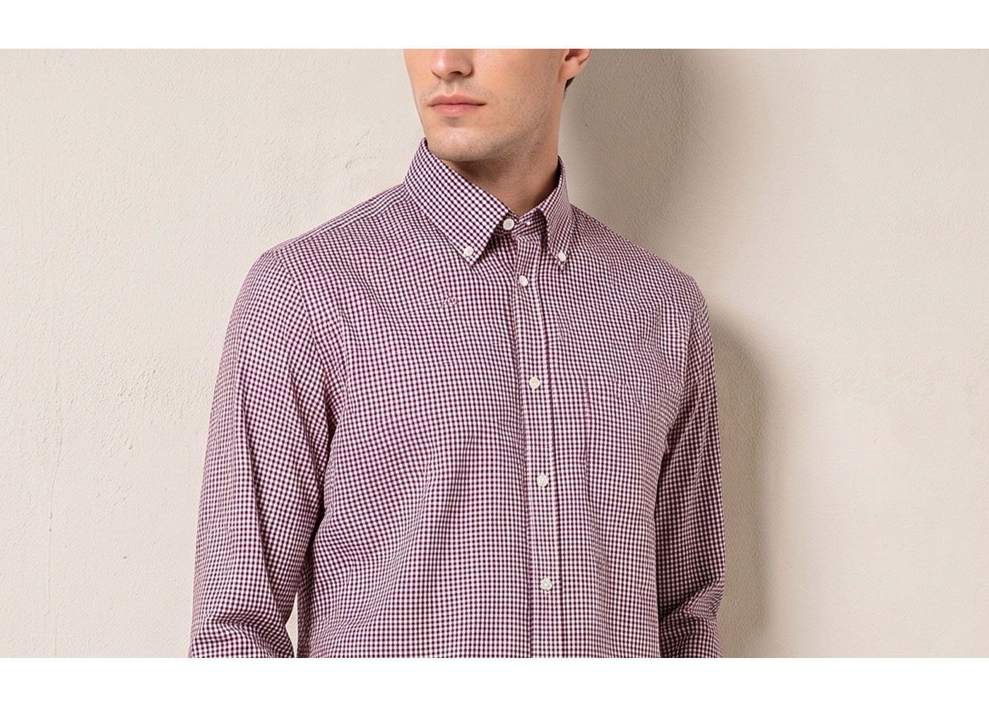Man wearing maroon and white gingham button-down long-sleeve shirt with buttoned collar and chest pocket against a pale wall