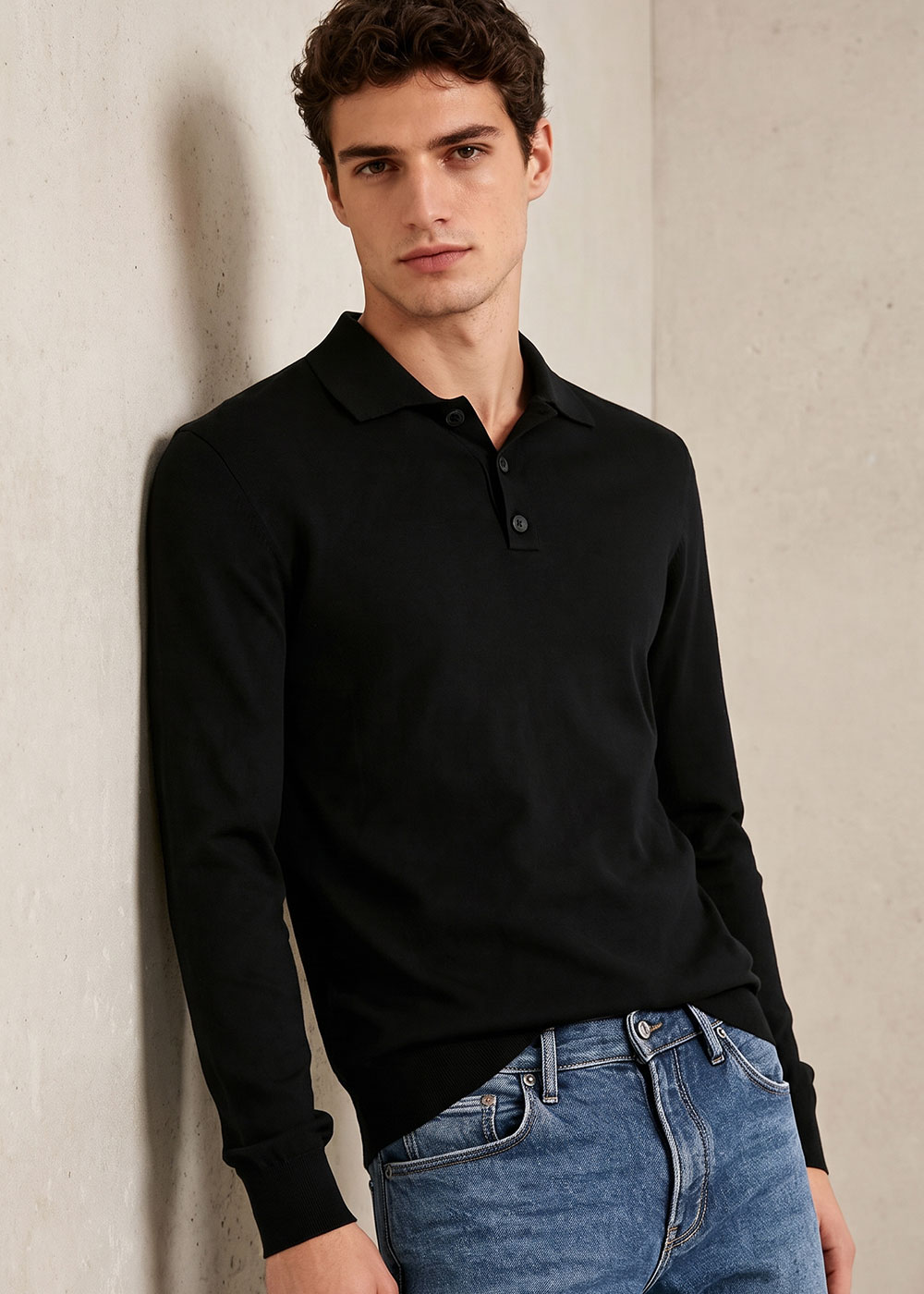 Young man leaning against concrete wall wearing black long-sleeve polo sweater and blue jeans, neutral expression