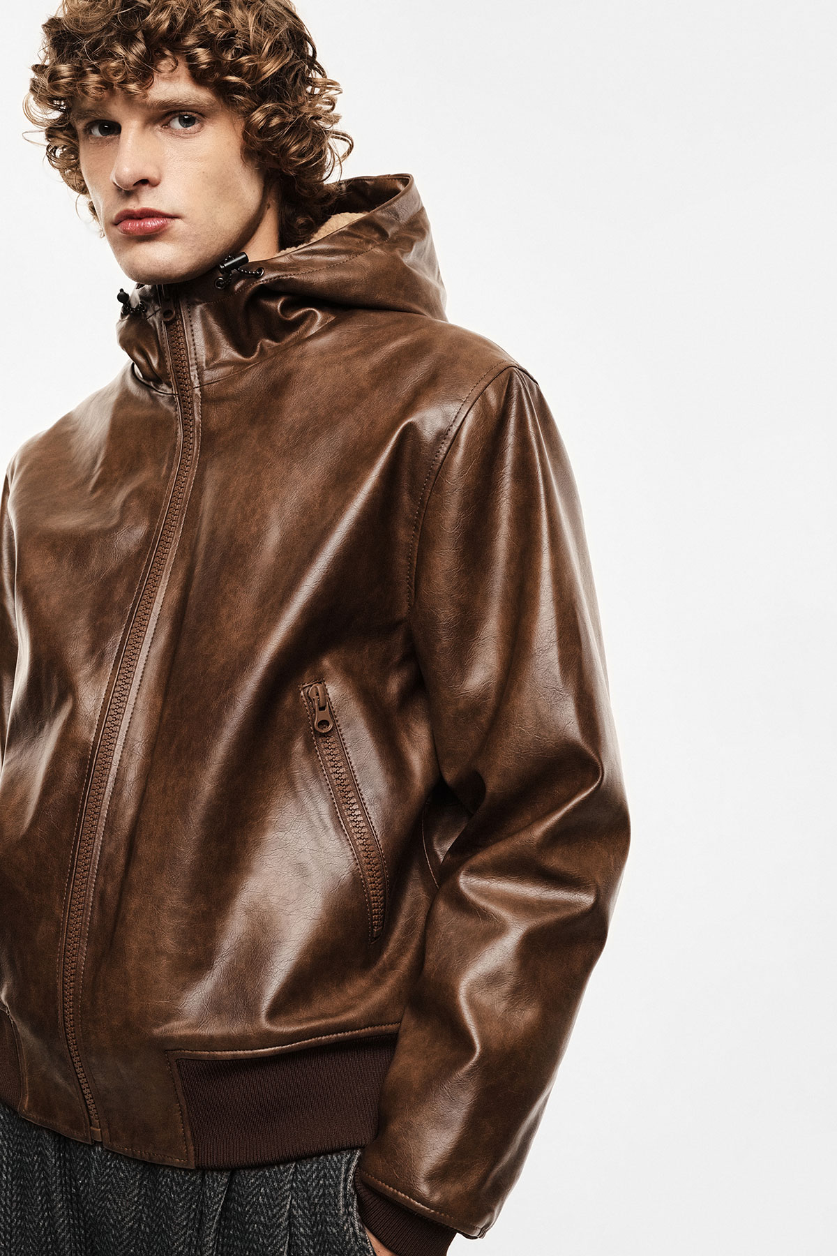 Young man modelling brown leather hooded jacket with zip front, side pockets and ribbed hem against pale background