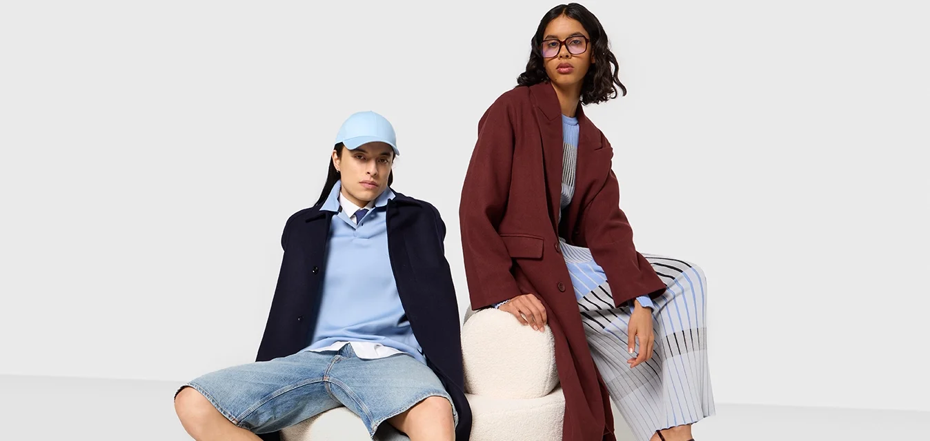Due modelli sono seduti su uno sgabello rotondo, di colore crema e texture. Uno indossa un cappotto blu navy, una camicia azzurra chiara, pantaloni corti di denim e stivali neri. L'altro indossa un cappotto bordeaux, un top azzurro chiaro, pantaloni a righe a gamba larga e scarpe platfform bordeaux.