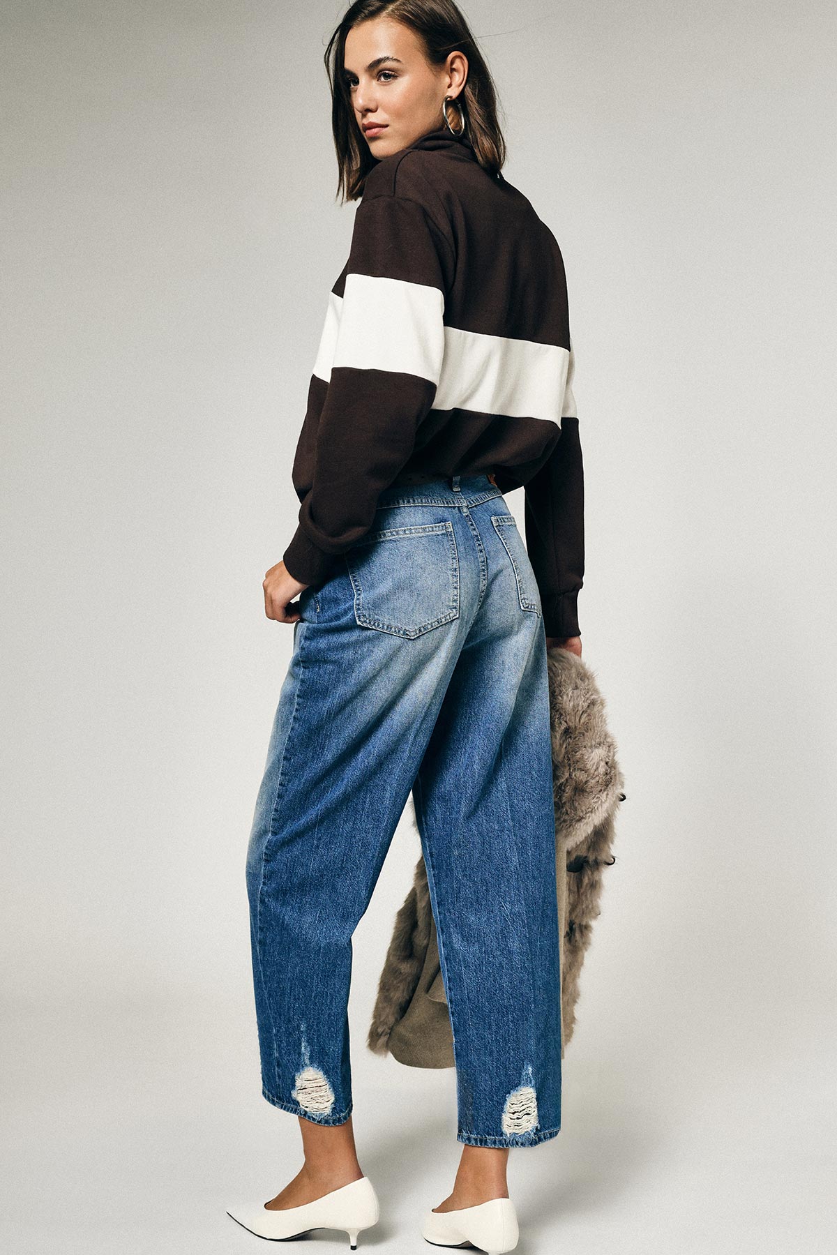 Woman seen over shoulder wearing brown sweatshirt with white stripe, high‑waist cropped distressed blue jeans, white heels and holding a faux‑fur coat