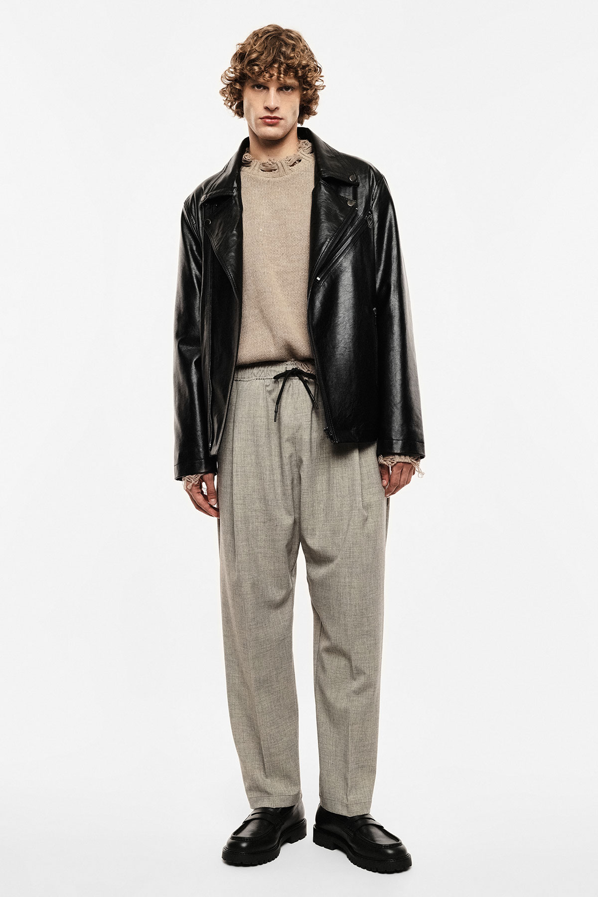 Male model wearing a black leather jacket, beige knit jumper, grey tailored drawstring trousers and black loafers on a white background