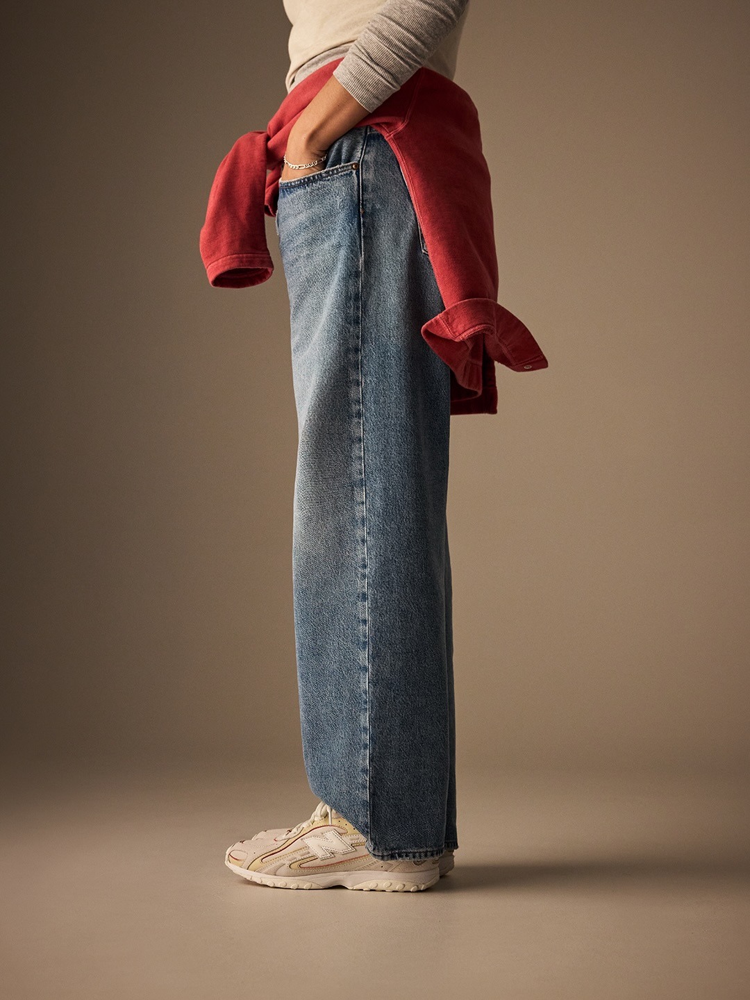 Side view of person wearing wide-leg blue jeans, white trainers and a red sweatshirt tied at the waist, hand in pocket