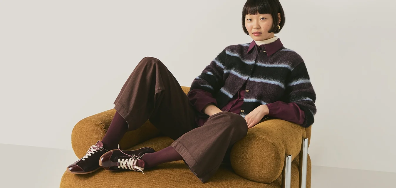 Donna con capelli corti e scuri seduta casualmente su una grande poltrona giallo senape, indossa un maglione a righe, pantaloni larghi e scarpe da ginnastica.