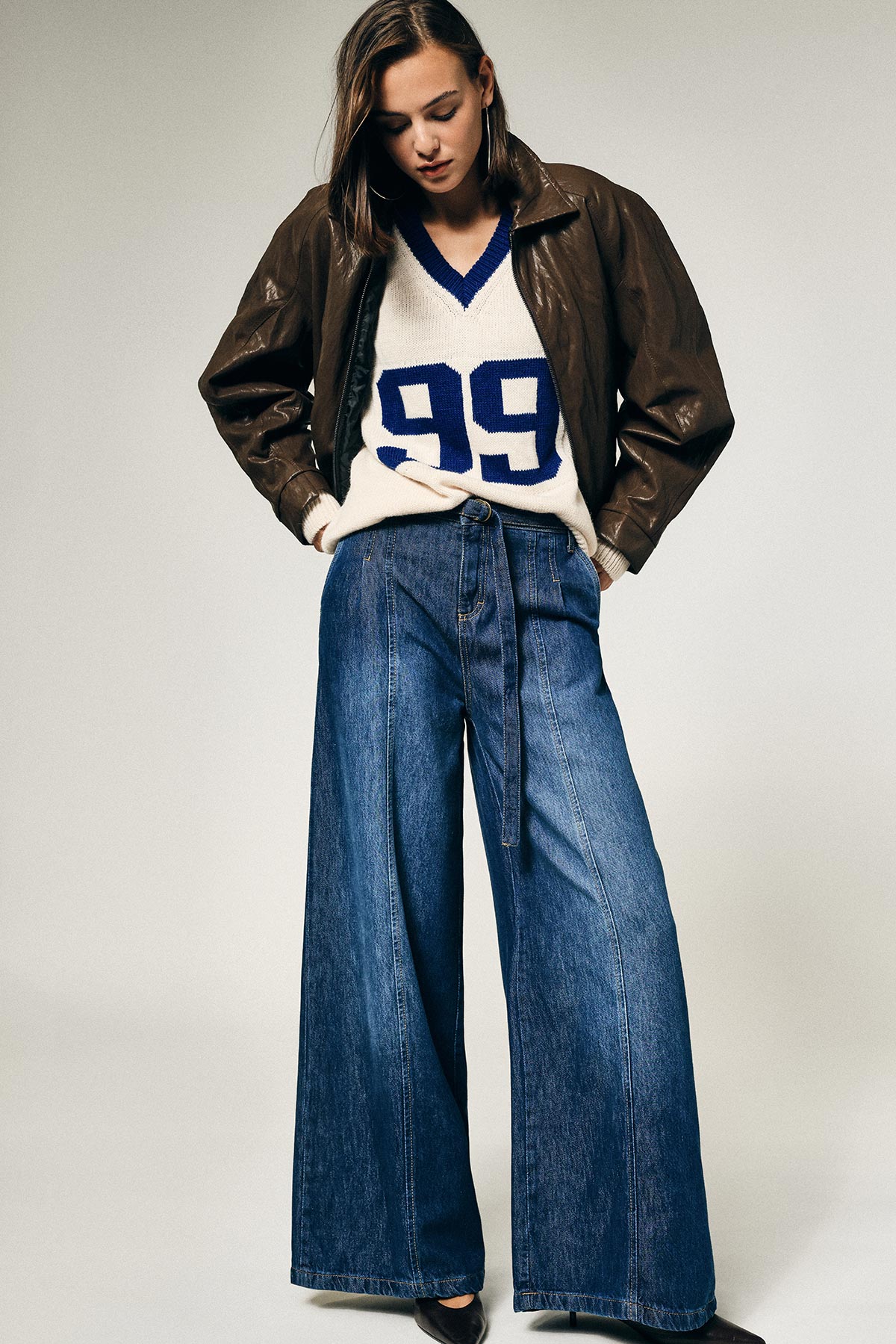 Young woman in brown leather jacket and cream sweater with blue '99', wide-leg denim jeans, hands in pockets, looking down