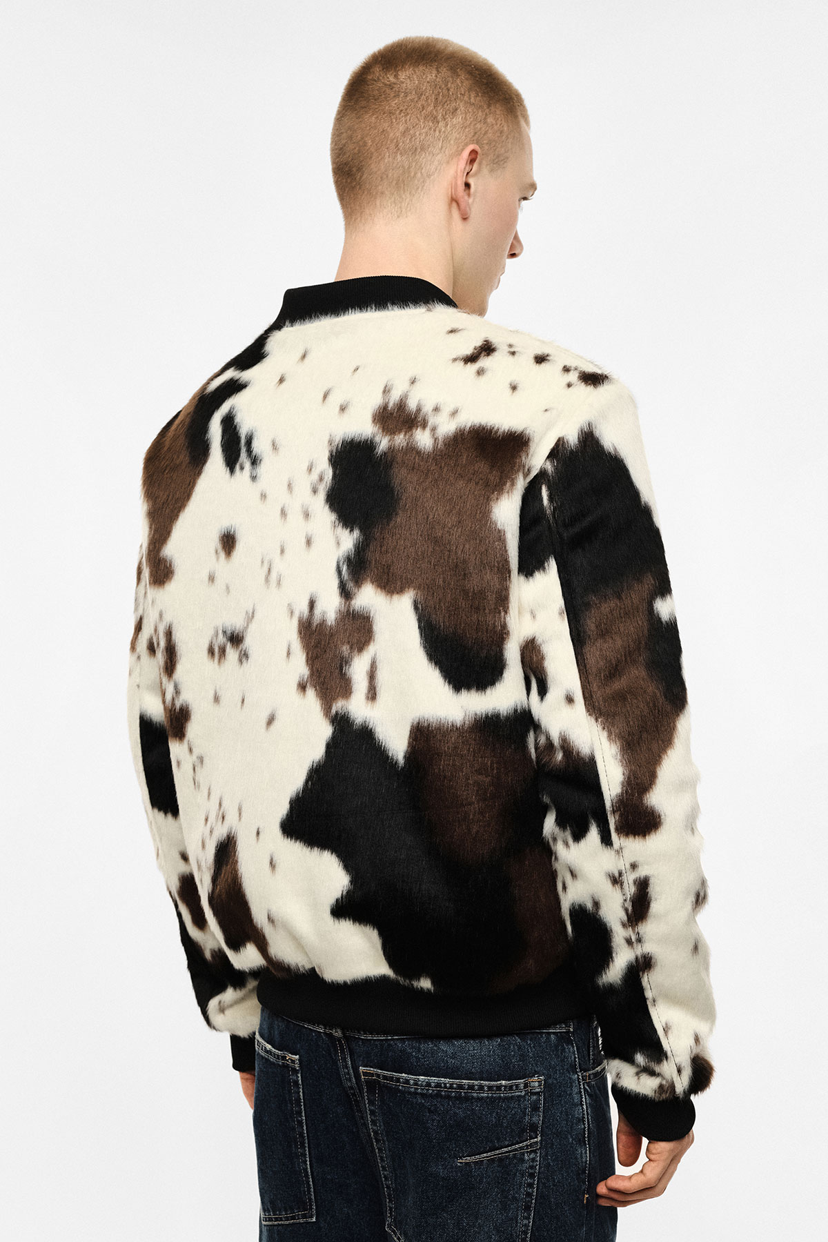 Back view of model wearing a black-and-white cow-print faux fur bomber jacket and denim jeans on a white background