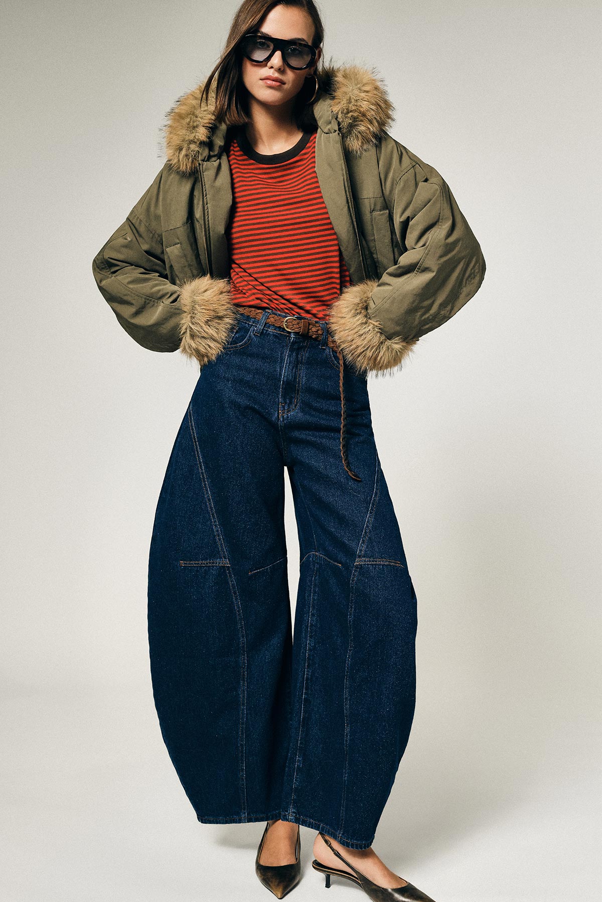 Model wearing olive fur-trim parka, red striped top, wide-leg denim jeans, sunglasses and slingback heels on plain background