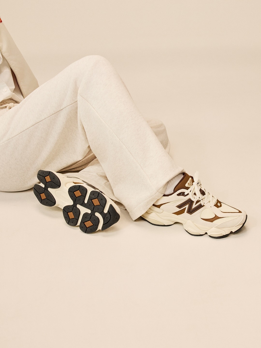 Person seated wearing cream and brown chunky trainers with patterned black soles and cream trousers on a beige background
