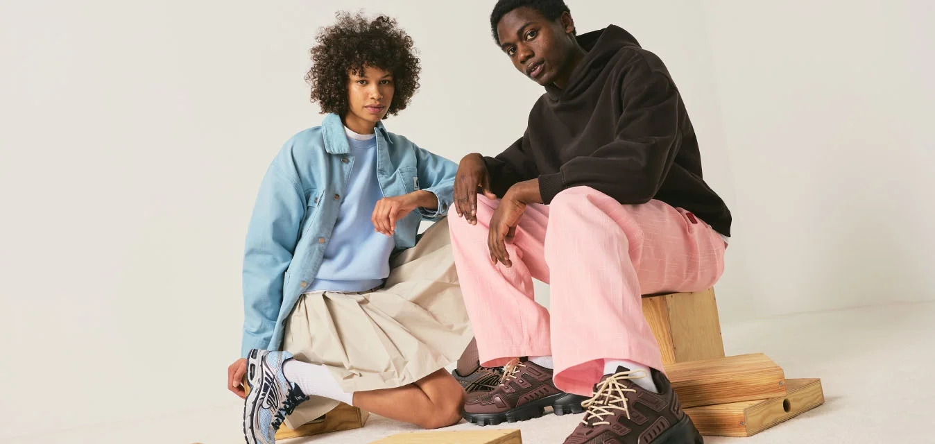 Due giovani seduti su e vicino a casse di legno su uno sfondo bianco uniforme, vestiti con abbigliamento casual da strada e sneakers.