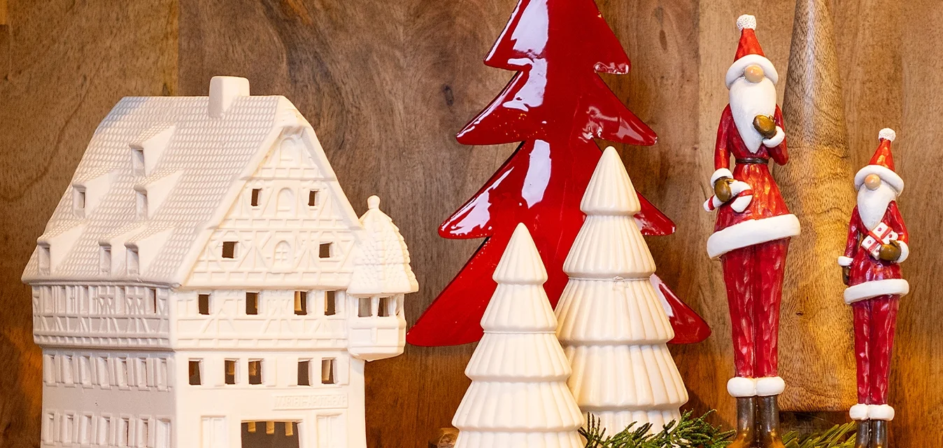 Casa in ceramica bianca con tetto dettagliato, albero lucido rosso, tre alberi conici bianchi e due figure di Babbo Natale rosse. Sfondo in legno naturale.