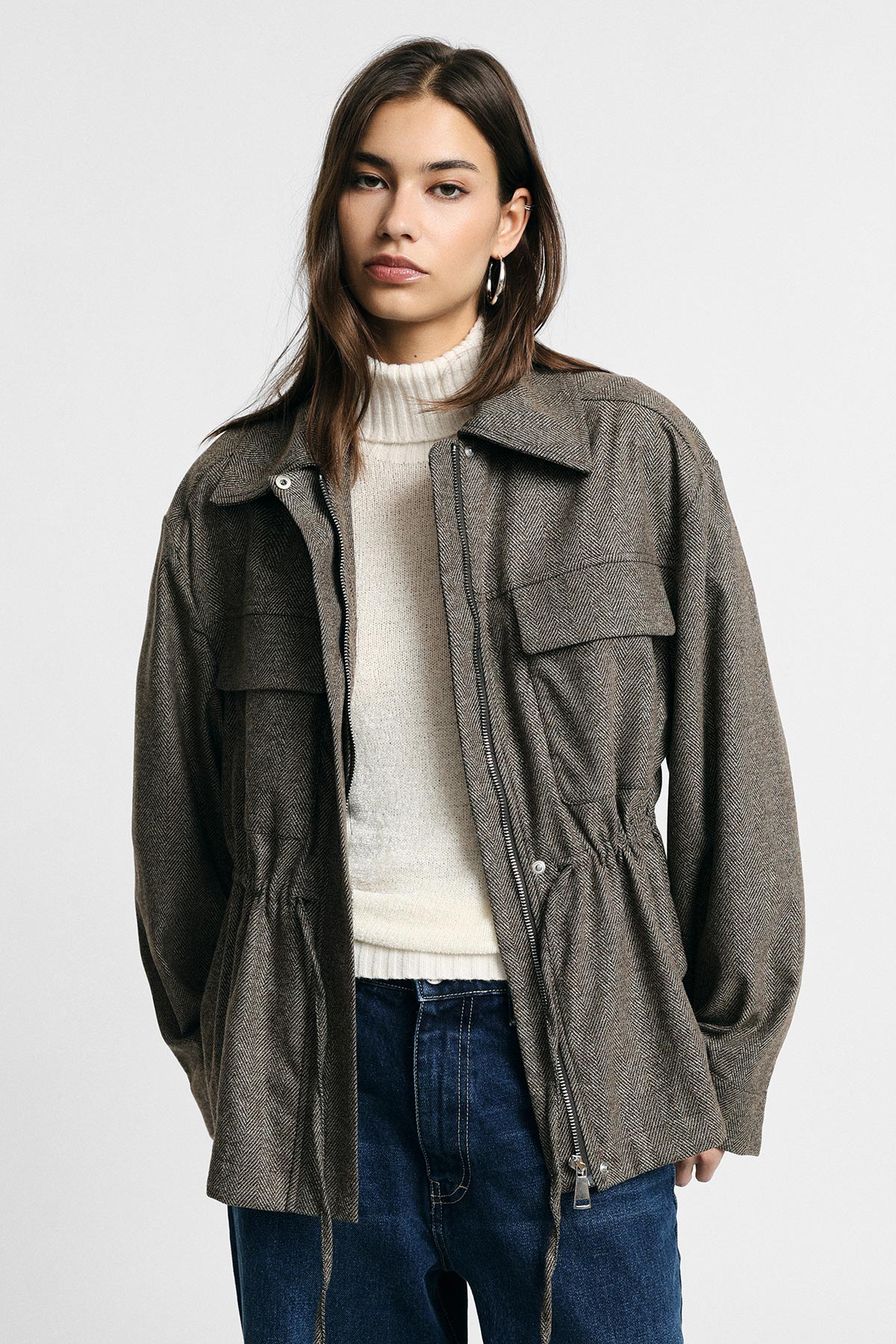 Young woman wearing a brown herringbone utility jacket over a cream roll-neck sweater and blue jeans against a plain background.