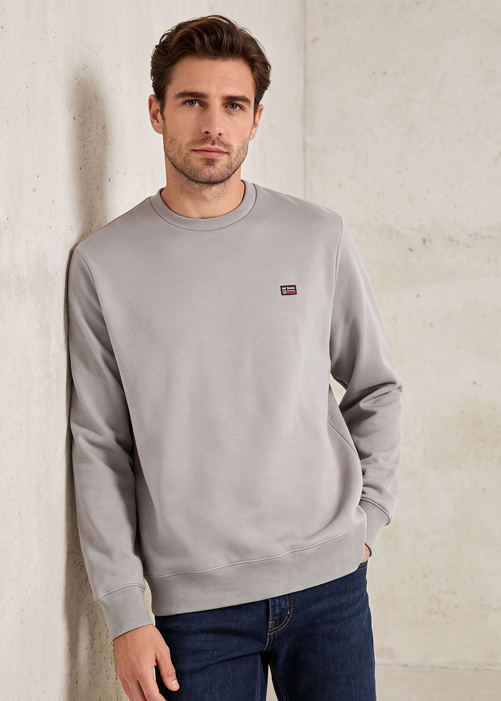 Male model in a light grey crewneck sweatshirt and dark jeans leaning against a concrete wall