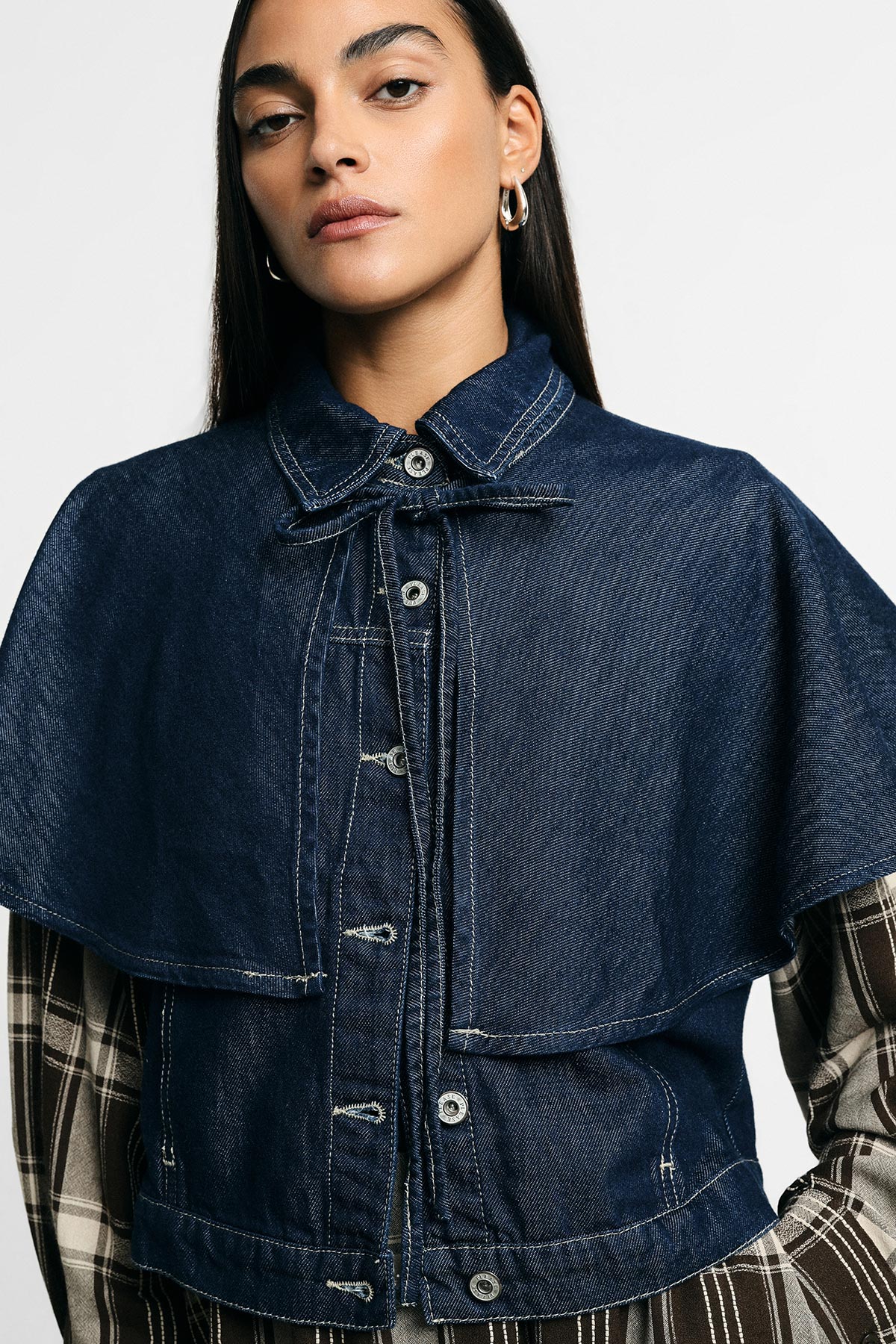 Woman model wearing cropped dark denim cape jacket with button front over plaid long sleeves, single hoop earring visible