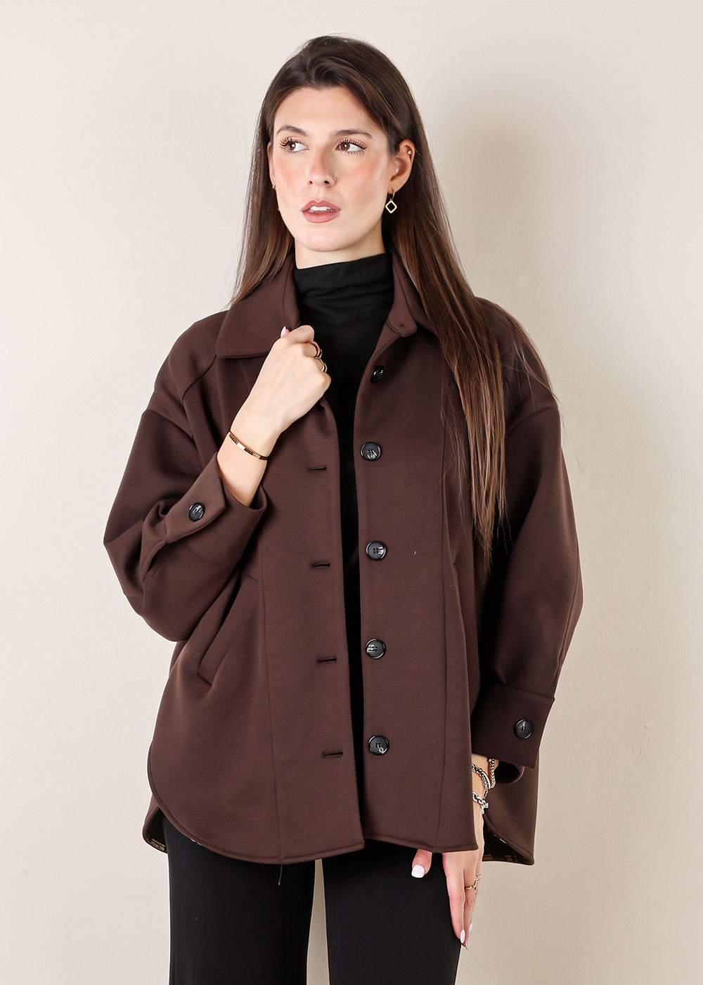 Woman wearing oversized brown button-up jacket over black top, long brown hair, gold jewellery, neutral background