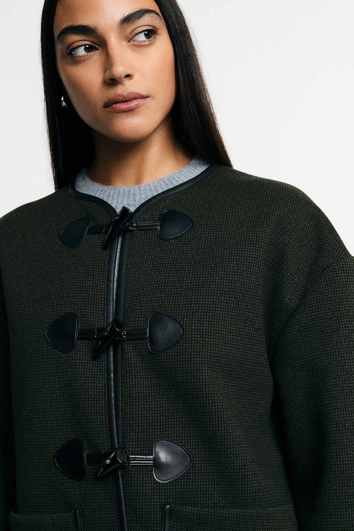 Woman in dark green textured toggle coat with black leather trim, looking to the side