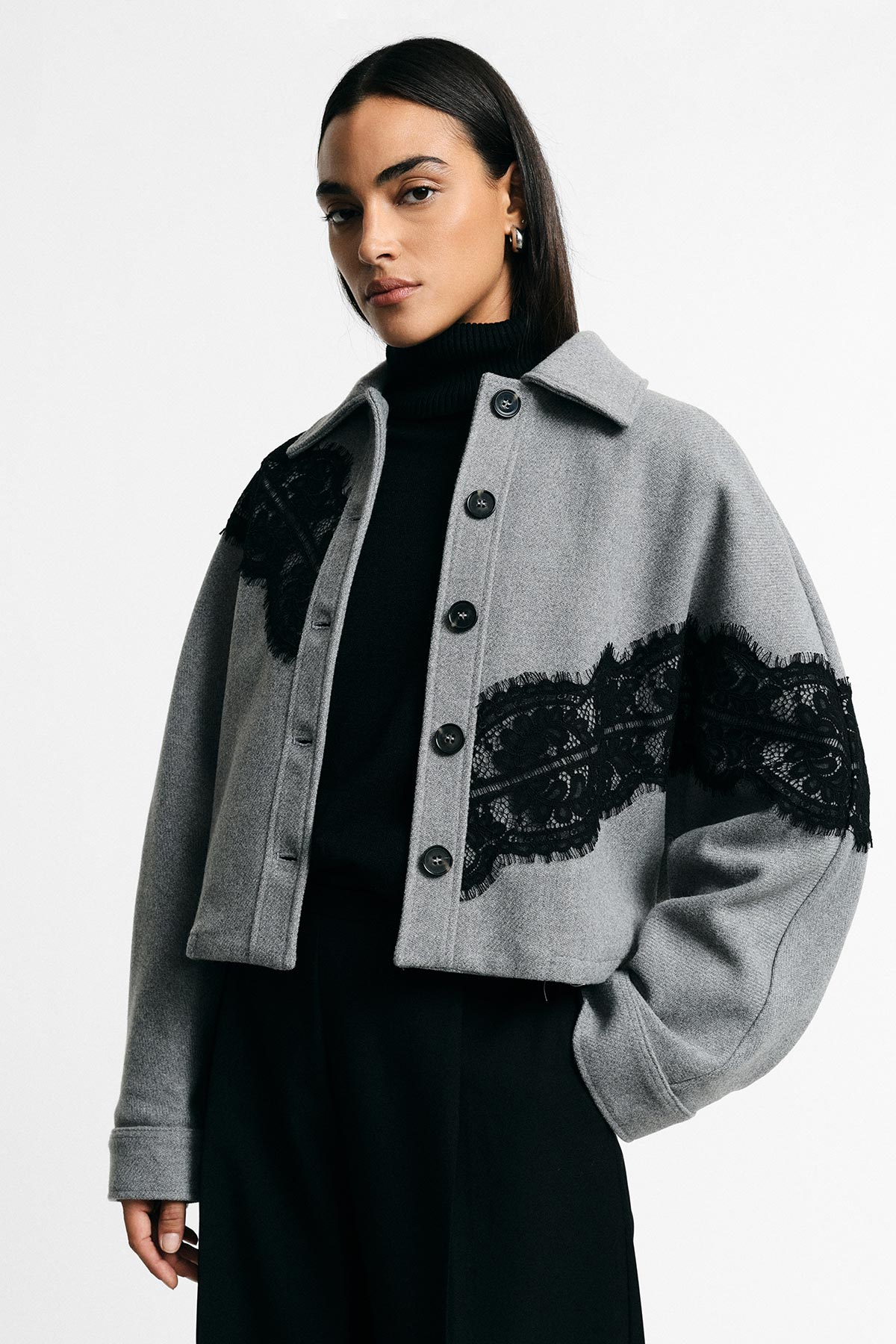 Woman wearing cropped grey jacket with black lace trim over a black roll-neck, hands in pockets, neutral studio background