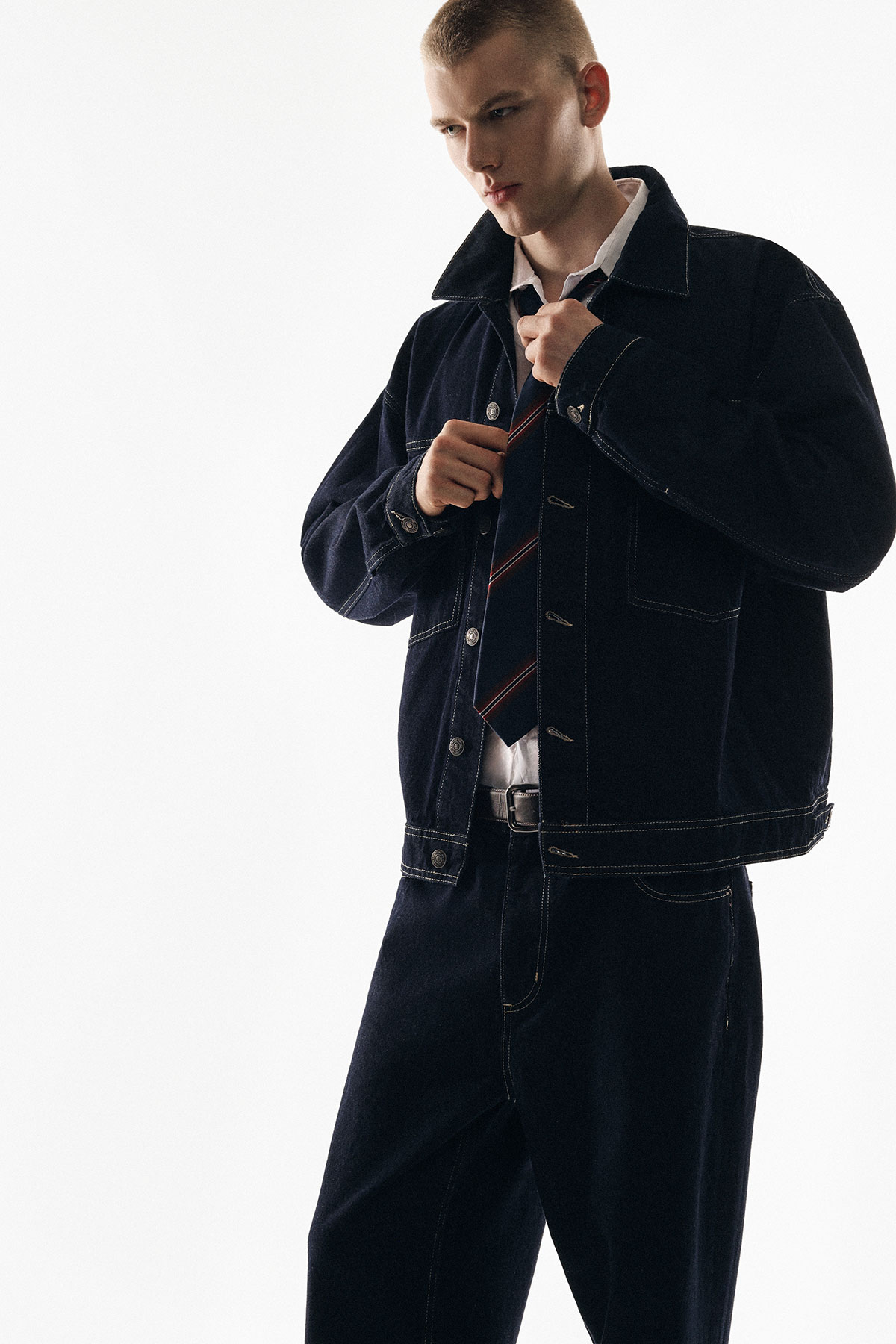Young man adjusting a striped tie while wearing a dark denim jacket and matching trousers against a plain white background