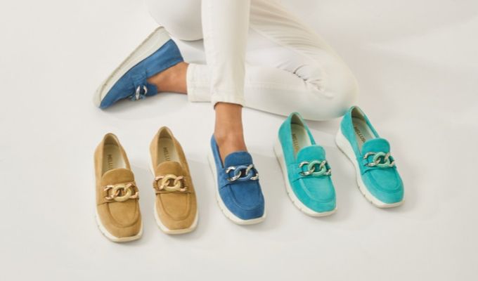Blue suede slip-on loafers with silver chain detail worn by model in white pants, shown alongside tan and turquoise pairs.