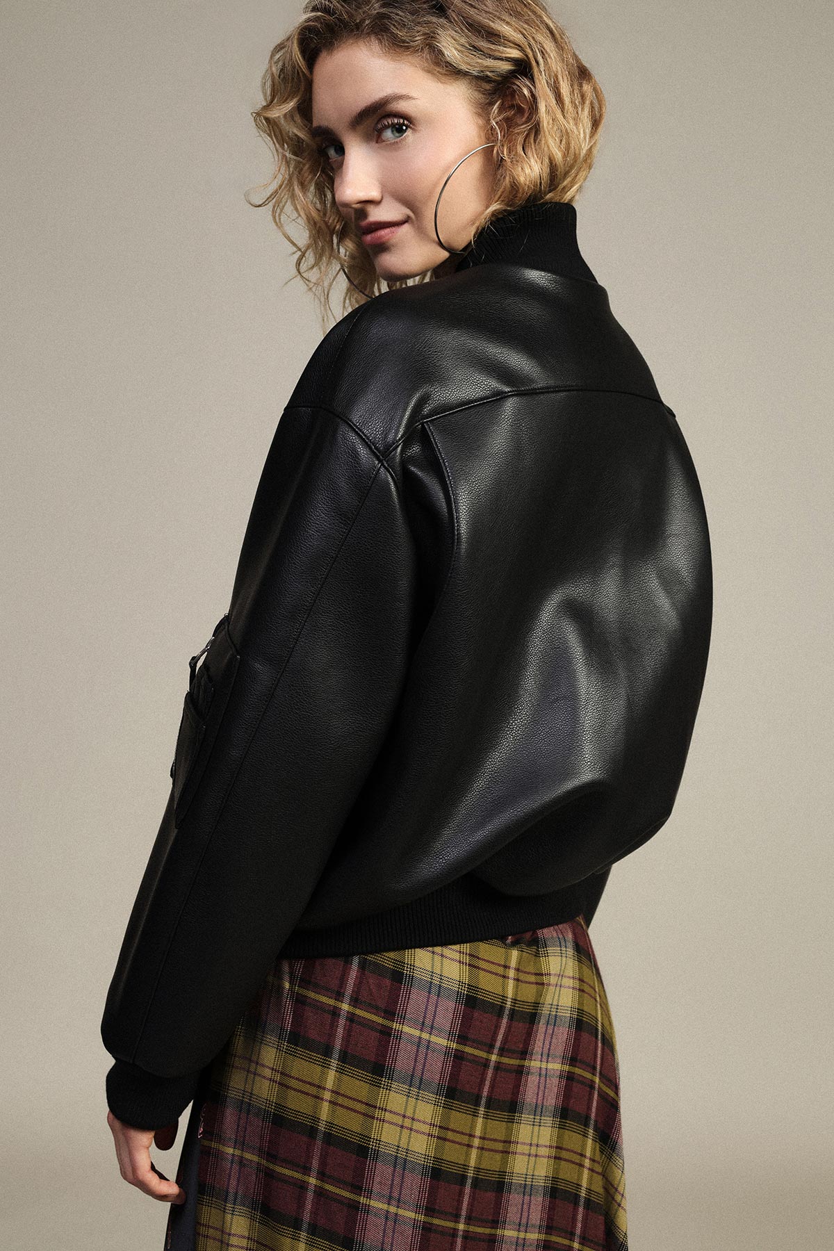 Woman in black leather bomber jacket and yellow plaid skirt, hoop earring, looking over shoulder, fashion portrait