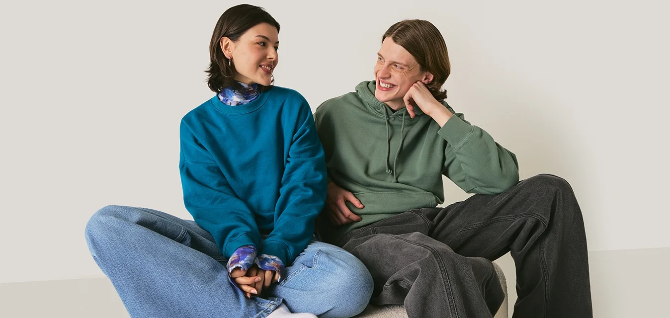 Due persone sedute su una superficie morbida. Una indossa una felpa blu petrolio e jeans chiari; l'altra indossa un hoodie verde e pantaloni grigio scuro.