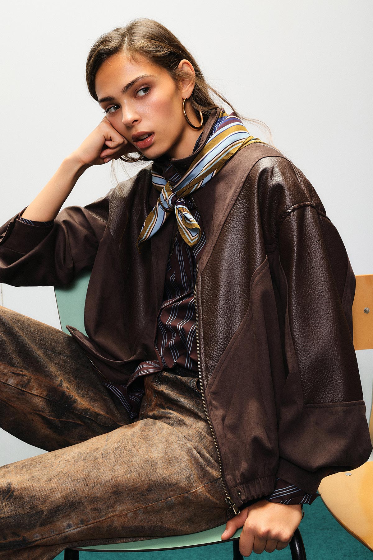 Young woman model seated wearing a brown leather jacket, striped shirt, patterned silk neck scarf and gold hoop earrings