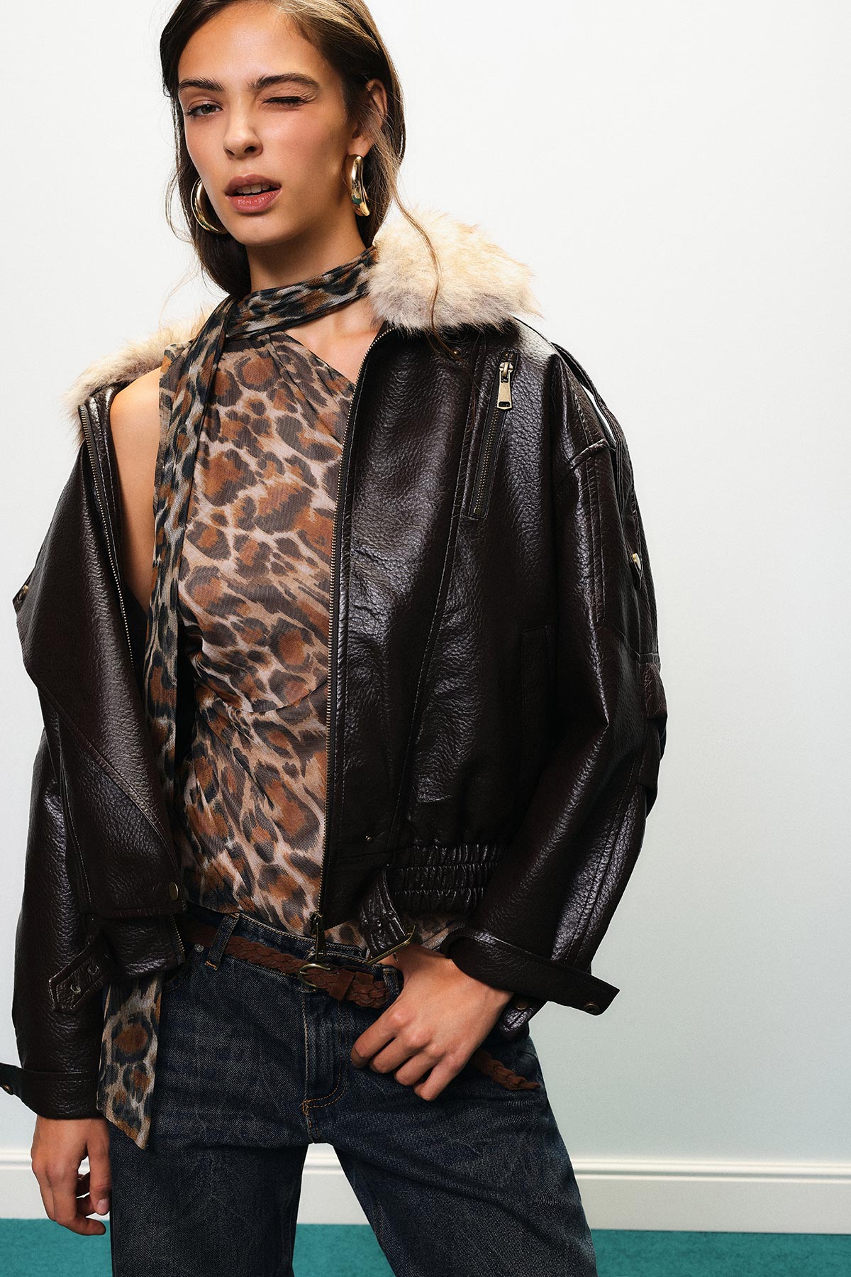 Woman winking in brown leather shearling jacket over leopard-print top, high-waist jeans and gold hoop earrings, hand on hip