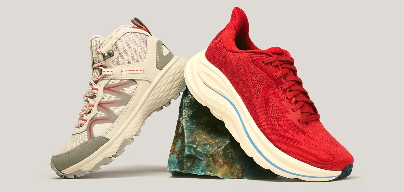 Scarpa da trekking beige e scarpa da corsa rossa bilanciate su una piccola roccia su uno sfondo semplice.