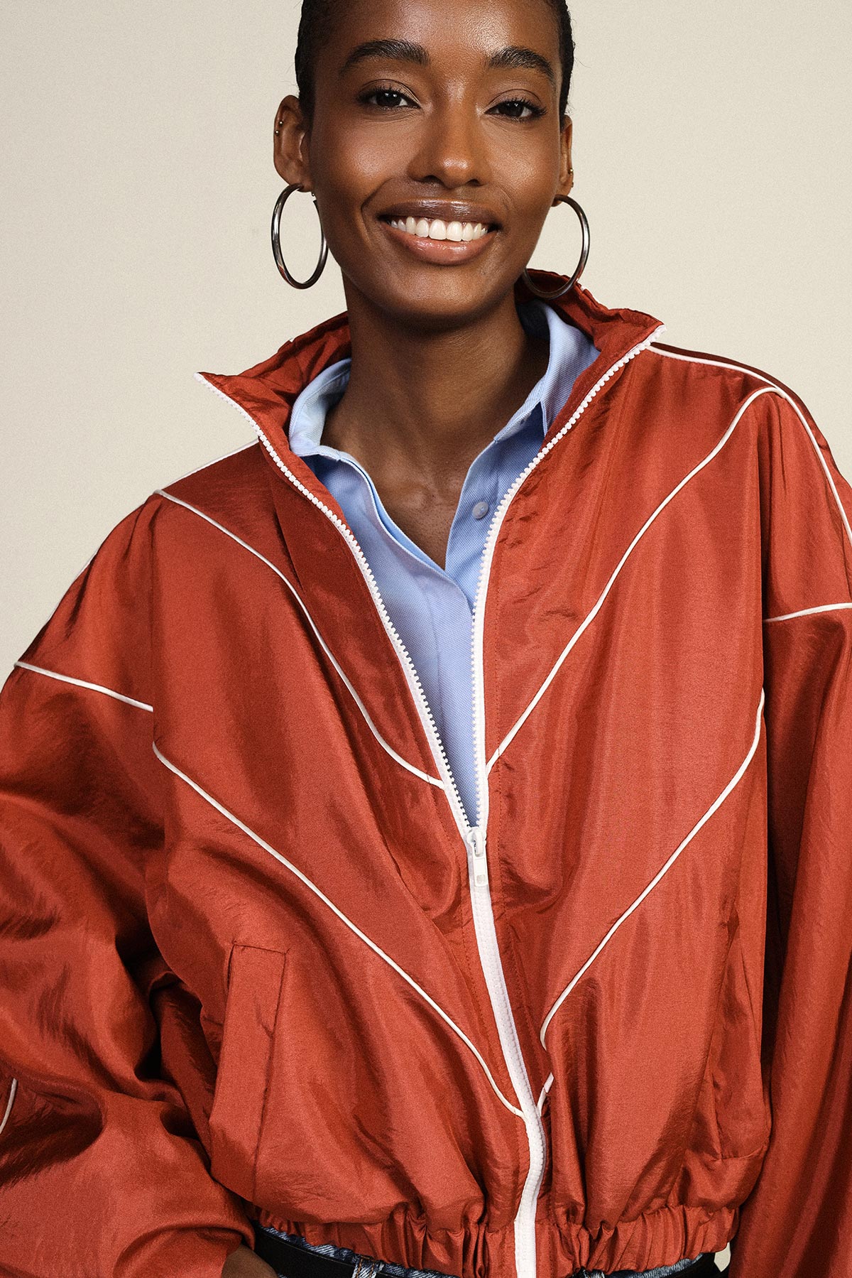 Smiling woman in a red zip-up windbreaker over a blue collared shirt, wearing large hoop earrings