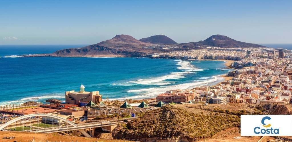 Aerial view of a coastal city with turquoise Atlantic sea, a long sandy beach, curved bridge, harbour buildings and volcanic hills